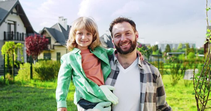 Portrait shot of happy Caucasian father holding cute little son on hands in green beautiful garden. Summertime work at house. Working in orchard with kid. Sunny day.