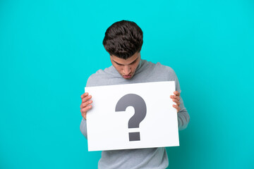 Young handsome caucasian man isolated on blue bakcground holding a placard with question mark symbol