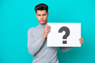 Young handsome caucasian man isolated on blue bakcground holding a placard with question mark symbol with sad expression