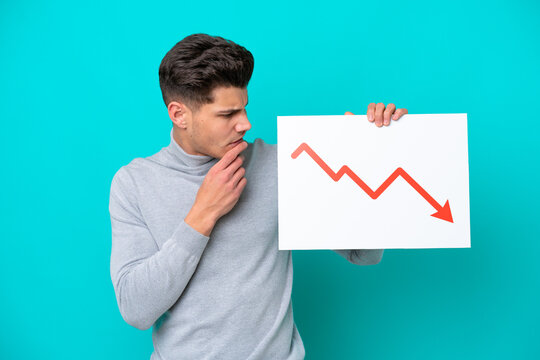 Young handsome caucasian man isolated on blue bakcground holding a sign with a decreasing statistics arrow symbol and thinking