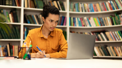 Focused woman using laptop and writing in notebook, studying online in library interior, panorama with free space