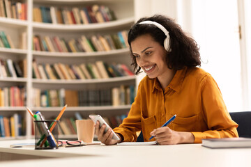 Happy female student sitting in library, using cellphone and taking notes while watching lesson,...