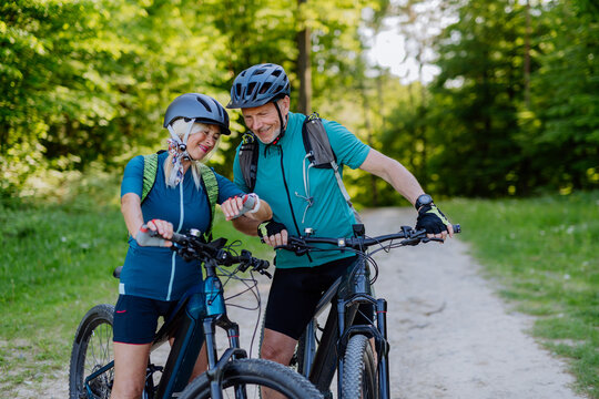 Portrait of active senior couple riding bicycles at summer park, looking at sports smartwatch, checking their performance.