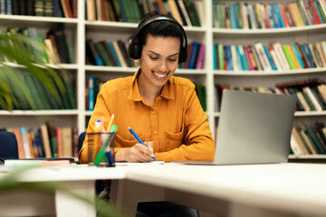 Online education concept. Smiling mixed race lady in headset studying remotely, sitting in library interior