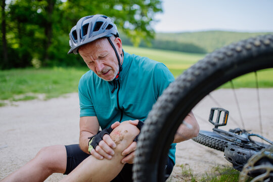 Active senior man in sportswear fell off bicycle on the ground and injured his knee, in park in summer.