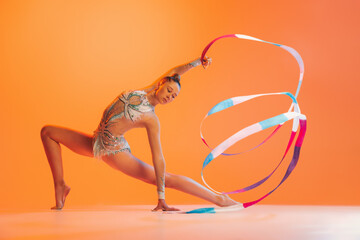 Young sportive girl, rhythmic gymnastics artist dancing isolated on orange color background. Concept of sport, action, aspiration, education, active lifestyle