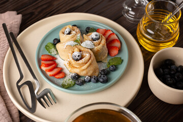 Russian thin pancakes, rolled into tubes, on a mint and beige plate on a dark background, strewn with blueberries with strawberries and mint, breakfast