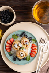 Russian thin pancakes, rolled into tubes, on a mint and beige plate on a dark background, strewn with blueberries with strawberries and mint, breakfast