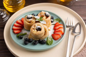 Russian thin pancakes, rolled into tubes, on a mint and beige plate on a dark background, strewn with blueberries with strawberries and mint, breakfast