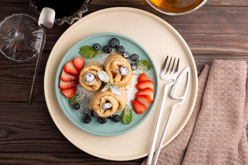 Russian thin pancakes, rolled into tubes, on a mint and beige plate on a dark background, strewn with blueberries with strawberries and mint, breakfast