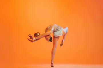 Zelfklevend Fotobehang One professional rhythmic gymnastics artist training with golden color ball isolated on orange background. Concept of sport, action, aspiration, competition © master1305