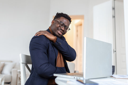 Tired African businessman working from home on his computer, feeling pain in his shoulder. man suffering from shoulder and back pain while sitting and working from home on laptop.