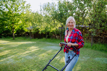 Elderly woman mowing grass with lawn mower in the garden, garden work concept.