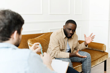 An irritated African American emotionally talks about problems to a psychologist at a session, a...