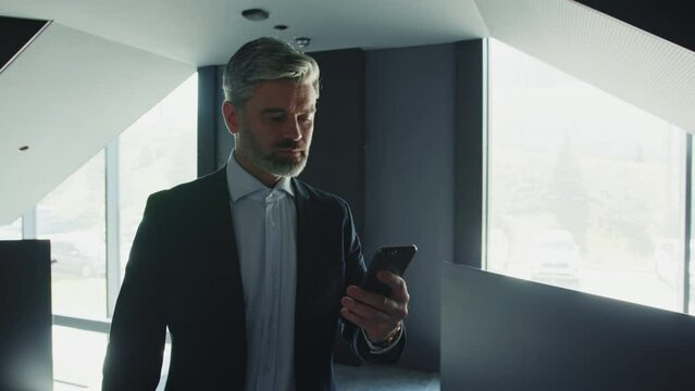 Shot of middle-aged gorgeous businessman with gray beard standing near stairs and using smrtphone. Serious man typing on phone in business center, holding mobile phone in hand, reading message