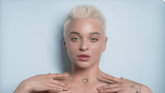 Young slim pretty Caucasian woman with white hair strokes her collarbones looking at the camera through rippling water on pale blue background | Flawless body concept