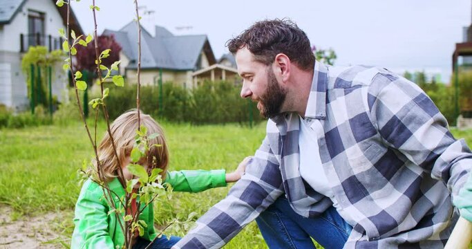 Happy cheerful small cute son sitting in garden with father at just planted little tree. Village house. Work at countryside. Boy with dad planting trees on sunny nice spring day.