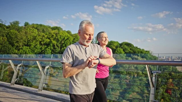 Active elderly man and woman jog on bridge against river