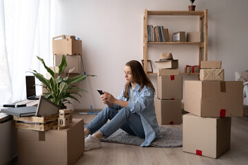 Attractive young woman is moving, sitting among cardboard boxes, using a smartphone, communicate...