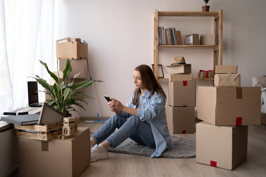 Attractive young woman is moving, sitting among cardboard boxes, using a smartphone, communicate via smartphone after moving to new flat.
