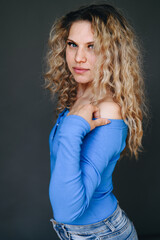 Vertical Portrait of Curly Blonde Woman in Blue Sweater Standing Sideways on Dark Background. Girl Without Makeup, Natural Appearance. Problem Skin Care Concept.