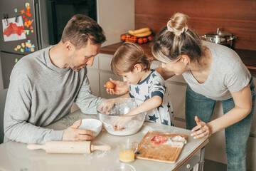 Cute boy passionate about kneading dough, holds egg in hand. Happy parents are watching him, teach baking. Family spends time in home kitchen. Real team