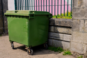 Green big size wheelie bin in a street. Garbage container ready for collection. Waste management industry in a city. Fence in the background. - obrazy, fototapety, plakaty