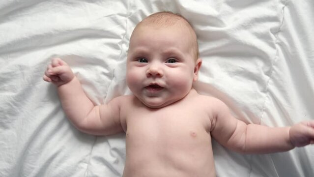 Happy naked baby in diaper lying on his back on the white sheet and touching his face. Baby girl with red hair looking at camera. Light background. Little child. Serious emotion. Advertising Mockup