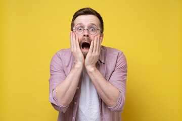 Man with glasses is shocked and frightened, he screams with mouth wide open, holds hands on cheeks...