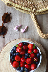 Round straw bag, brown sunglasses, pink earphones and bowl of berries on wooden table. Flat lay.