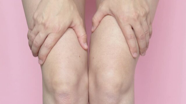 close up 4k video,woman is massaging with both hands her legs and knees.dry irritated skin.body care concept.pink background,isolated.
