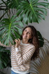 In harmony with nature. Portrait of young happy positive Italian woman touching Monstera leaf and smiling at camera, spending time with plants at home garden, enjoying gardening activity and plantcare