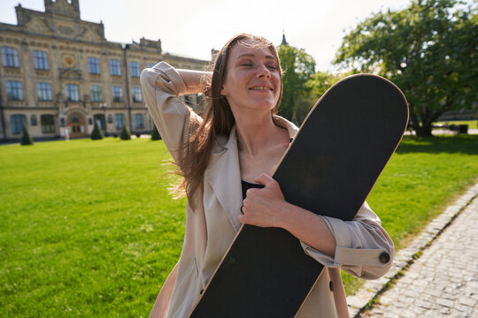 Lady with closed eyes stands in park with skateboard in hands