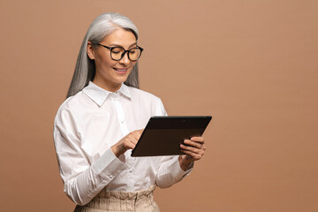 Modern elderly senior woman in formal wear using digital tablet isolated on beige. Portrait of...