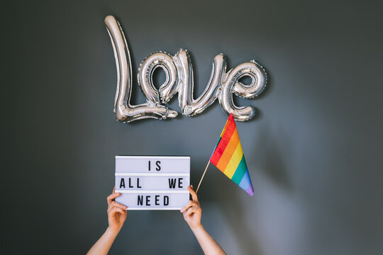 LGBTQ pride month background. Male hands holding lightbox with Text Love is all we need under silver foil balloon with rainbow flag on gray wall background. Film effect tonning. Selective focus.