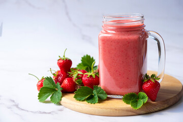 Strawberry smoothie or milkshake on wooden board with fresh strawberry fruit and green leaves on marble background. Strawberry drink in jar mug full of vitamins and antioxidants.
