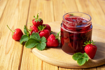 Strawberry jam in glass jar on wooden board with fresh strawberry fruit and green leaves on wooden background. Recipe of delicious homemade berry jam of strawberry full of vitamins and antioxidants.