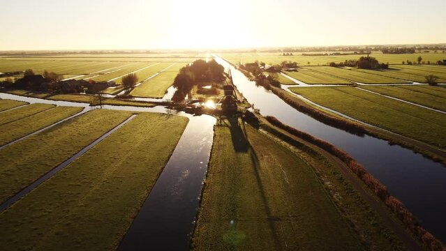 Aerial drone footage of vast green farmland fields in the Netherlands, separated by water canals with a windmill in the middle. Autumn sunrise.