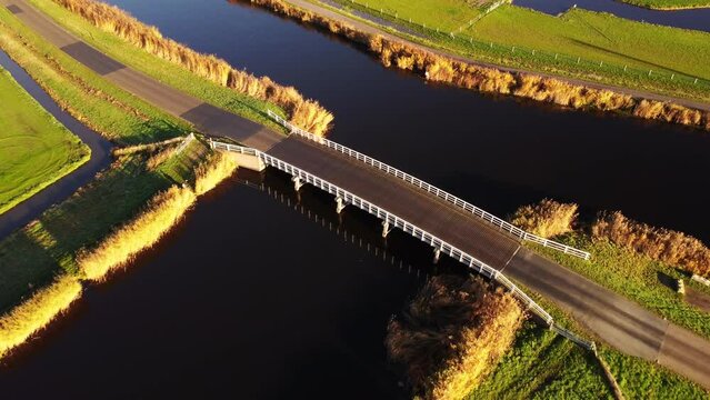 Aerial drone footage of a small road bridge among vast green farmland fields in the Dutch countryside, separated by water canals. Filmed at sunrise.