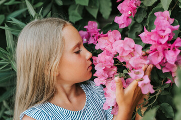 Fototapeta premium portrait face of candid little kid girl of eight years old on background of green plants and admired enjoy smell pink flowers during a summer vacation travel. gen z mental health concept