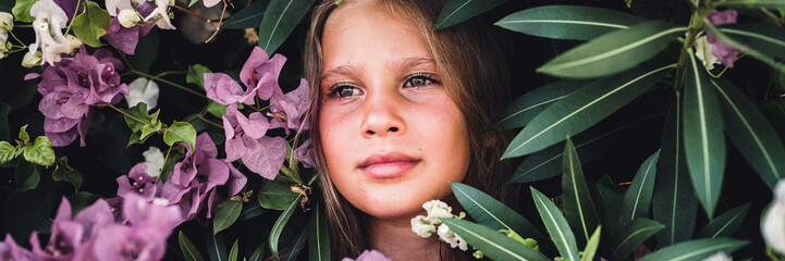 portrait face of candid beautiful little kid girl of eight years old with brown eyes on background of green plants and pink flowers during a summer vacation travel. gen z mental health concept. banner