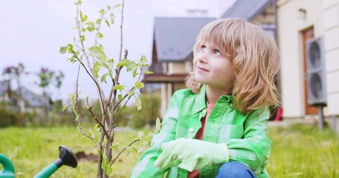 Portrait of cute little Caucasian boy sitting in garden at just planted tree in garden and looking at father with smile. Watering and planting trees in orchard. Spring day. Close up.