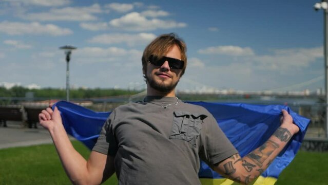 Happy caucasian man proudly representing his home country and putting Ukrainian flag on his back. Stylish Ukrainian dude wearing blue and yellow flag in slow motion outdoors as sign of patriotism
