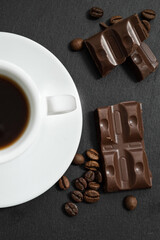 Top view on chocolate pieces and roasteed coffee beans scattered near white cup of coffee on dark background