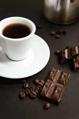 Chocolate pieces and roasteed coffee beans scattered near white cup of coffee on dark background
