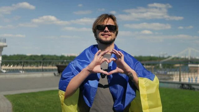 Long-haired guy in black shades wearing blue and yellow flag showing heart symbol with hands. Ukrainian man indicating that he supports love and positivity by performing heart gesture in slow motion
