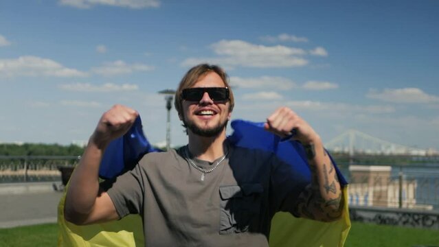 Happy smiling Ukrainian with flag of his country jumping and celebrating success and victory. Male caucasian model in sunglasses wearing blue and yellow banner expressing joy, triumph and delight
