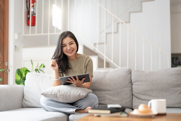 Beautiful attractive young Asian woman sitting on gray sofa and using digital tablet at living room in the morning.