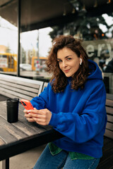 Trendy caucasian young woman using smartphone while sitting outdoor on summer terrace with smartphone. Curly girl wears blue pullover and jeans. Concept of electronic devices