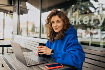 Lively young caucasian girl drinking coffee in morning and using her laptop. Brunette woman in casual clothes sits on summer terrace. Free time concept